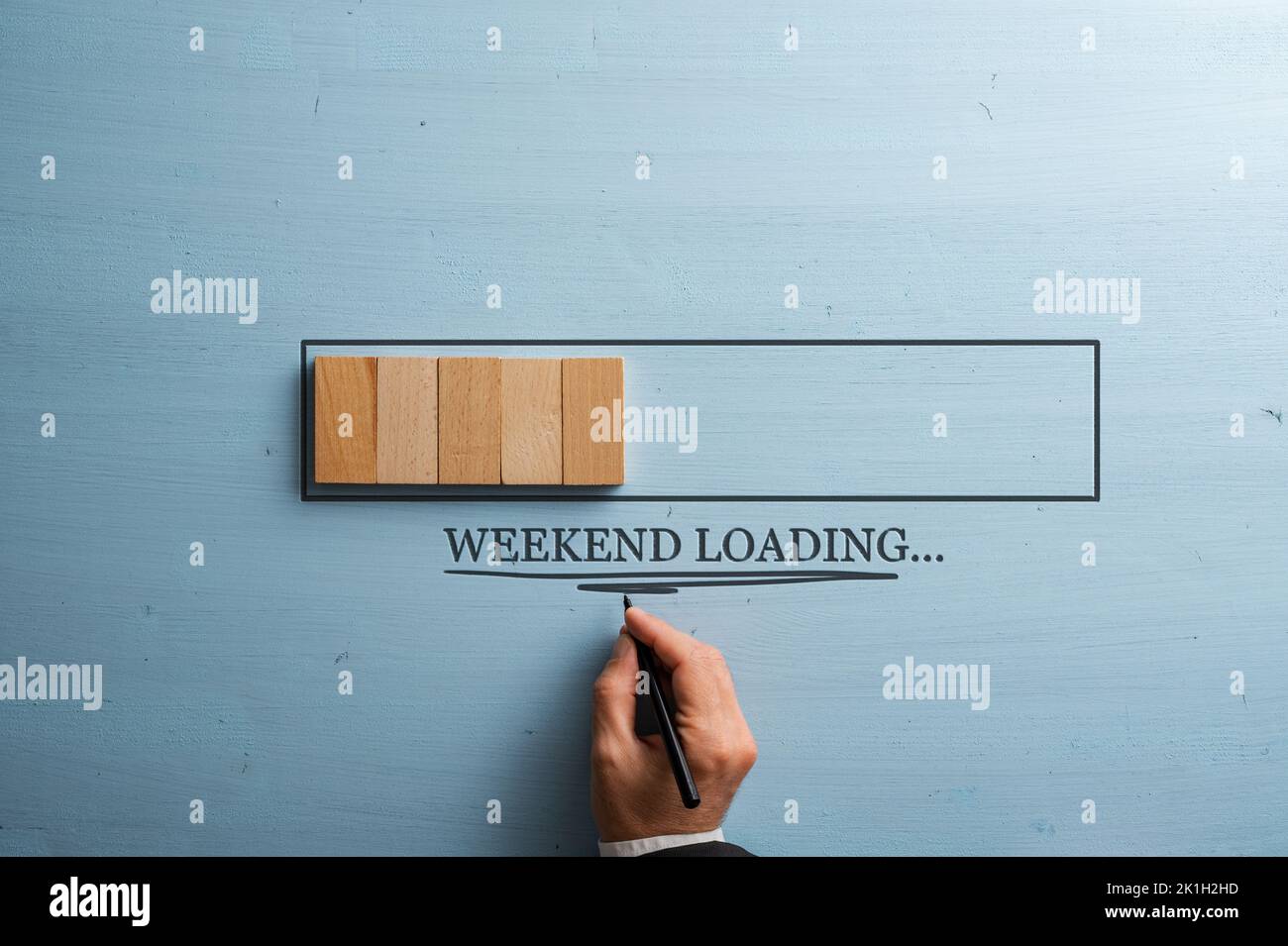 Male hand writing a Weekend loading sign under a loading bar made of wooden dominos. Over pastel blue wooden background. Stock Photo