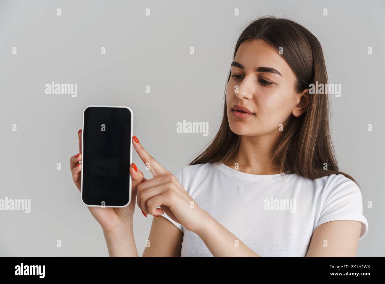 Portrait of a young casual white woman in t-shirt with long brunette hair standing over gray wall background showing blank screen mobile phone Stock Photo
