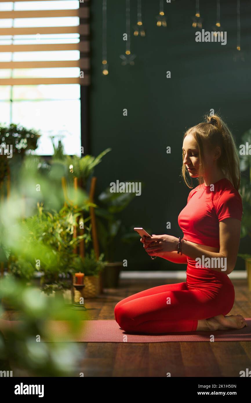 young female in red fitness clothes with smartphone doing yoga in the modern green house. Stock Photo