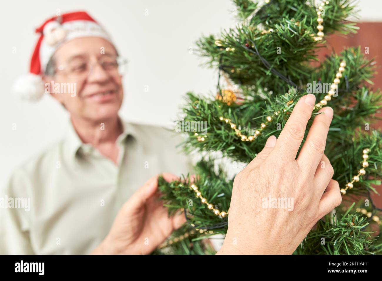 Senior hispanic couple decorating a Christmas tree, enjoying the holidays together. Female hands arranging the decorations in focus in the foreground, Stock Photo