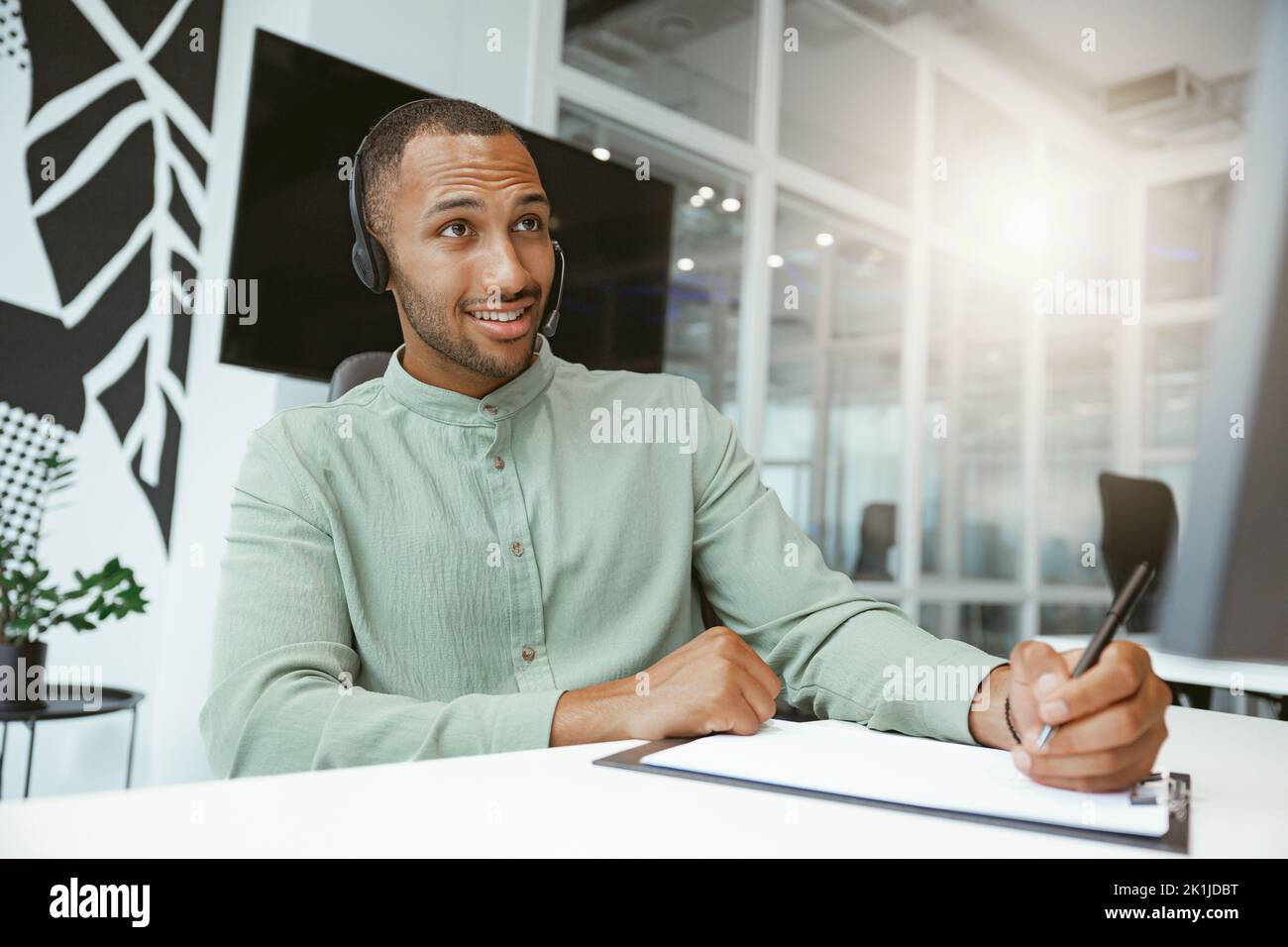 Confident African businessman taking notes while talking on phone with client in modern office Stock Photo