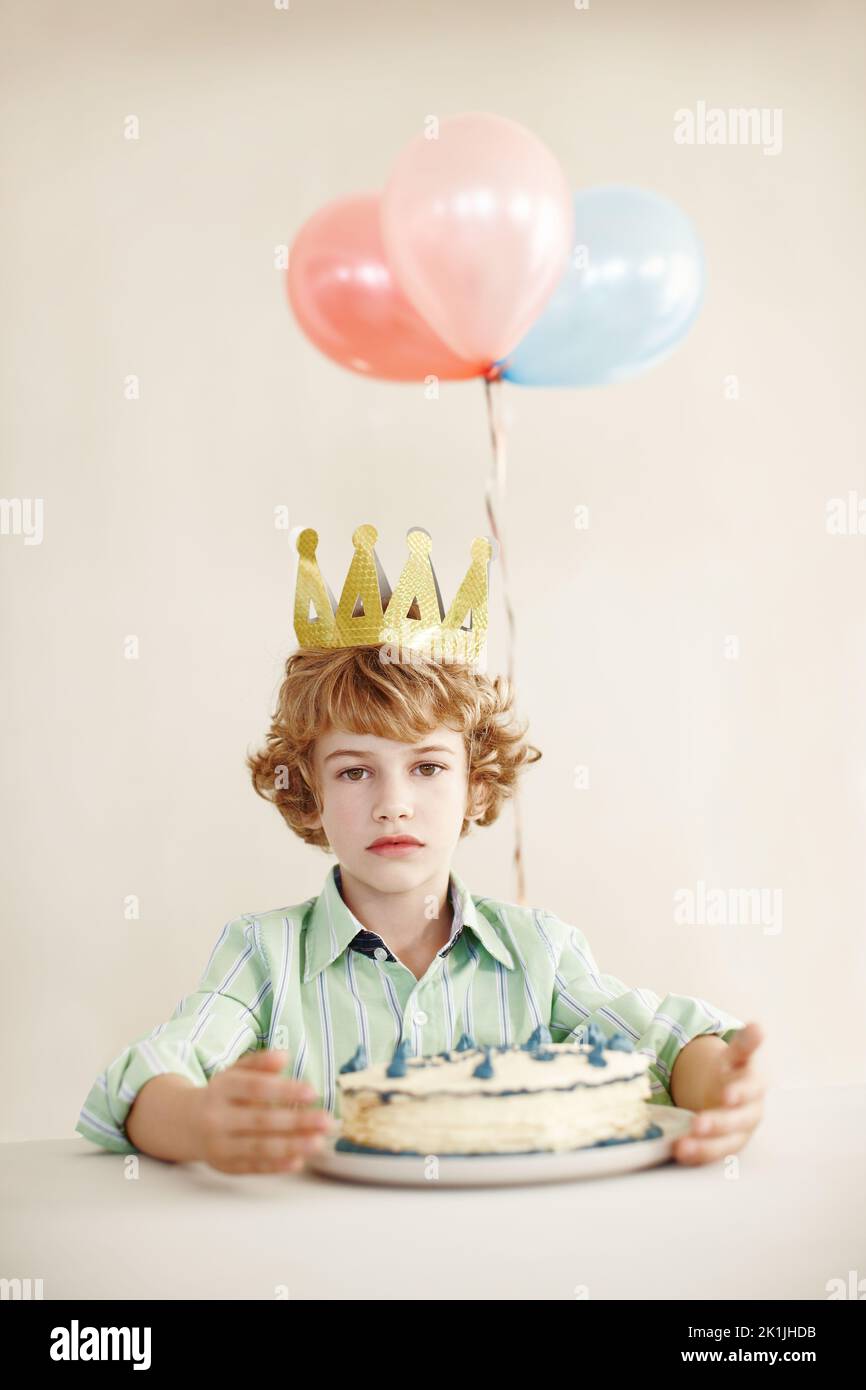 The king demands your presence. a cute little boy looking serious while sitting at a table with his birthday cake. Stock Photo