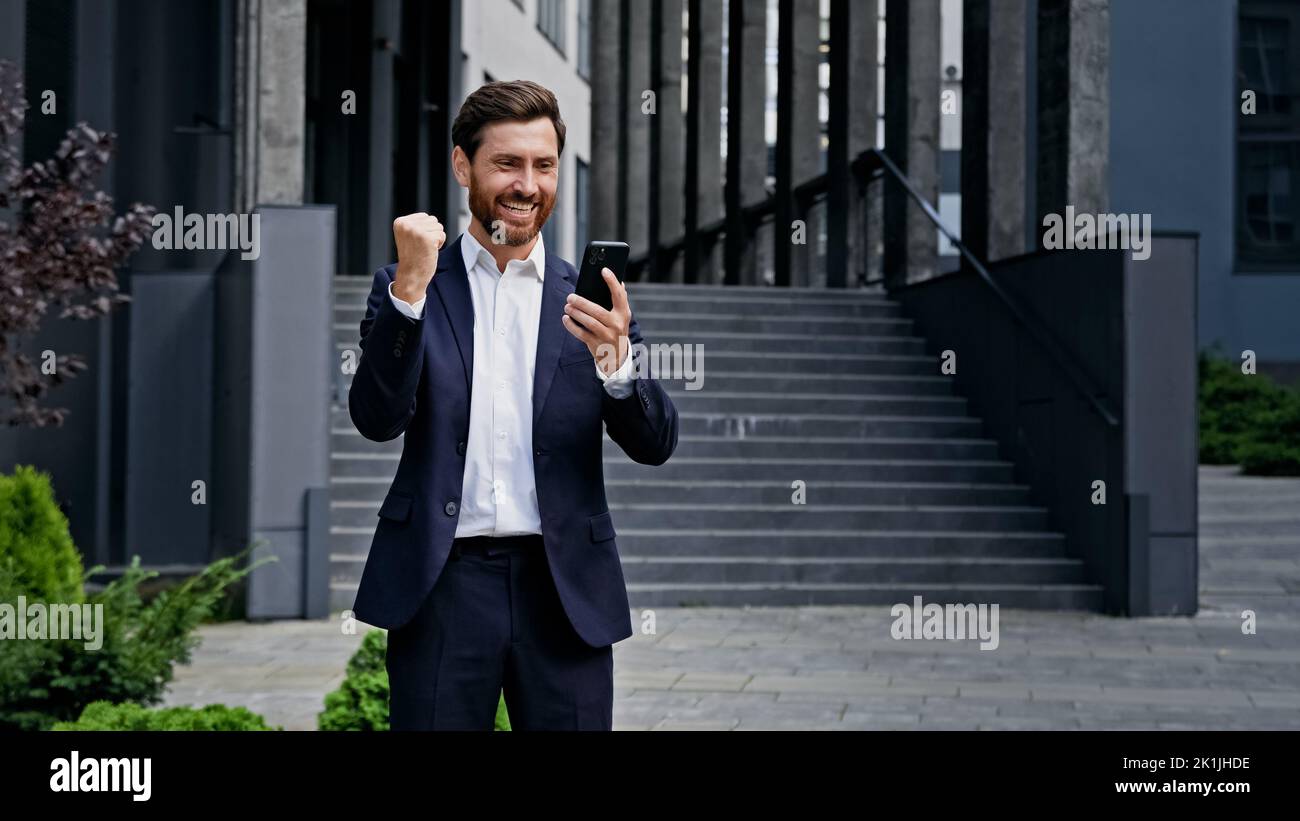 Excited enthusiastic young man standing outdoors reading email on phone with good news receiving great offer job promotion winning lottery receives Stock Photo