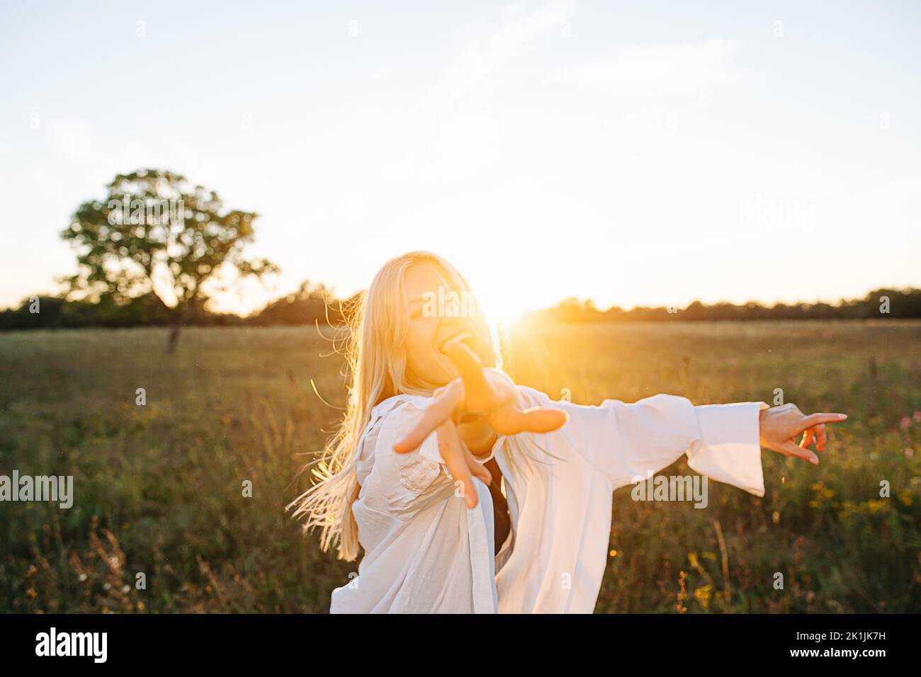 Excited young blond woman standing amidst wheat field. Reaching with her hand. Against setting sun, lit with soft bright orange light. Low angle. Stock Photo