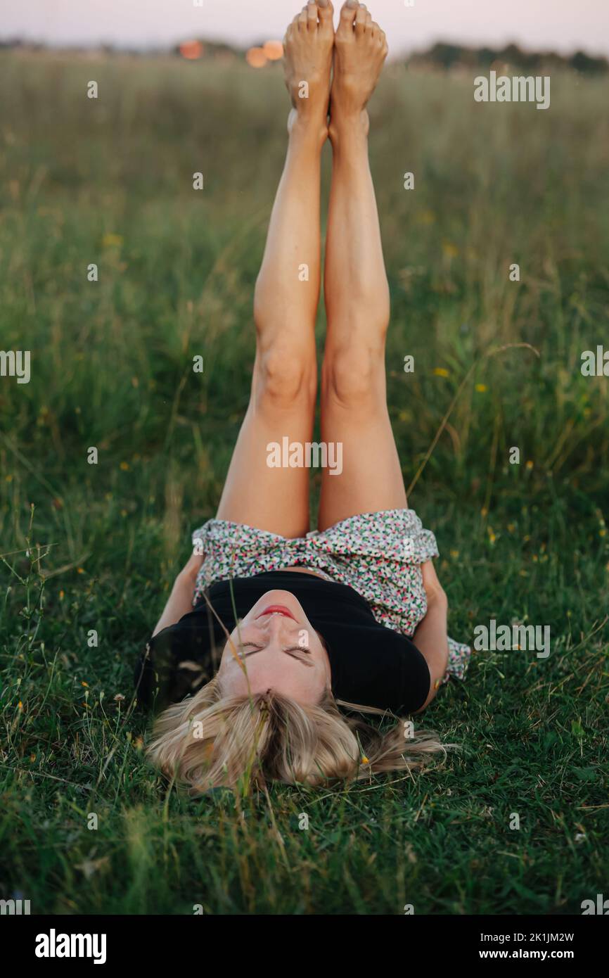 Exercising young blond woman holding her legs straight lying on her back. in a grass field . She is wearing skirt pants and black shirt. Stock Photo