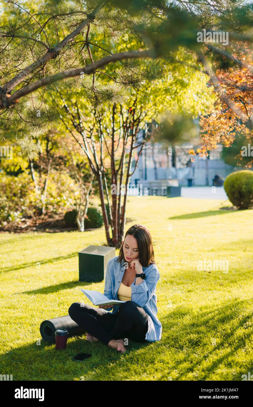 Pretty relaxed woman reading short story, lovely moments, sitting on green grass near yoga mat and mobile phone. Sport leisure concept. Harmony Stock Photo