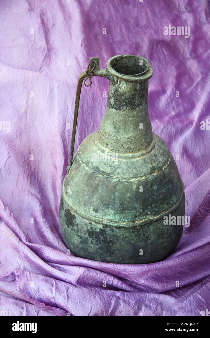 Still life with an antique Moroccan bronze jug on a purple fabric. Vertical photo with vintage jug on  lilac cloth Stock Photo