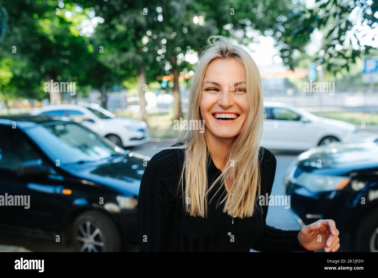 Positive young blond woman smiling in front of a car parking in background. Looking to the side. Stock Photo