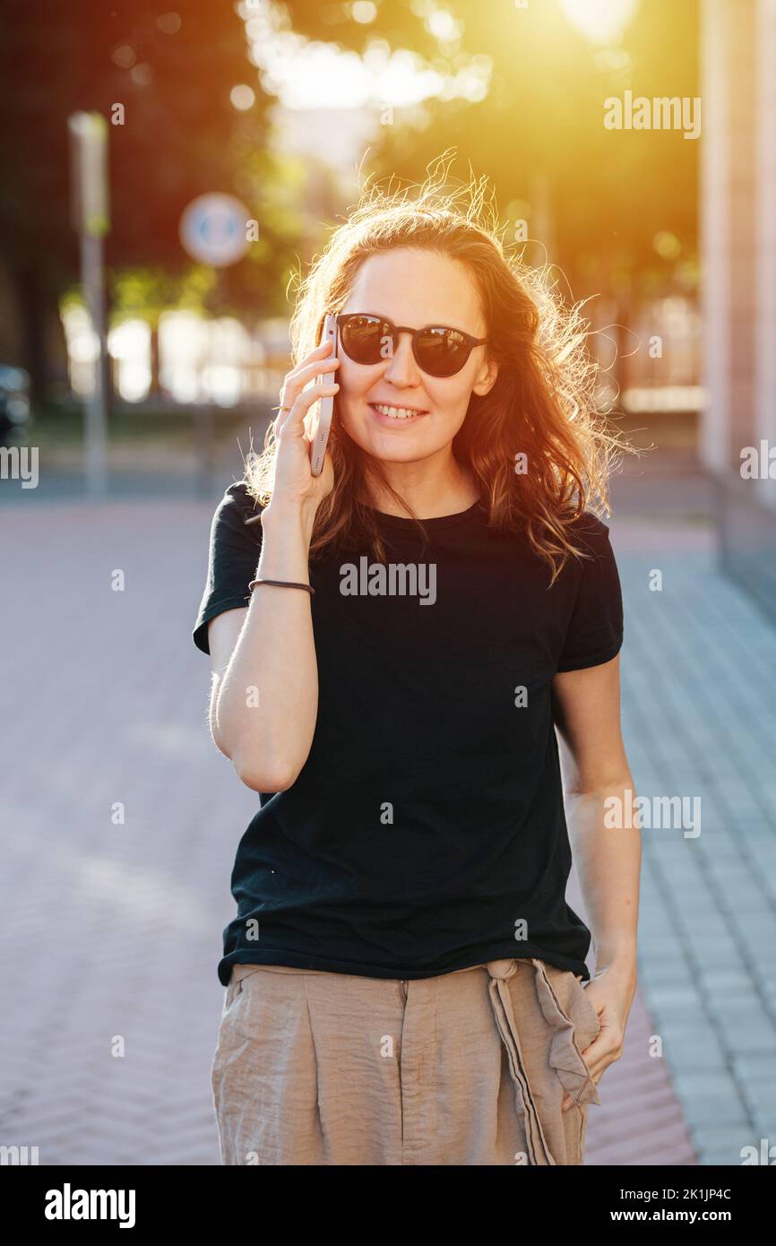 Smiling young woman in sunglasses happily chatting on her phone while walking on a pavement on a sunlit street. Stock Photo