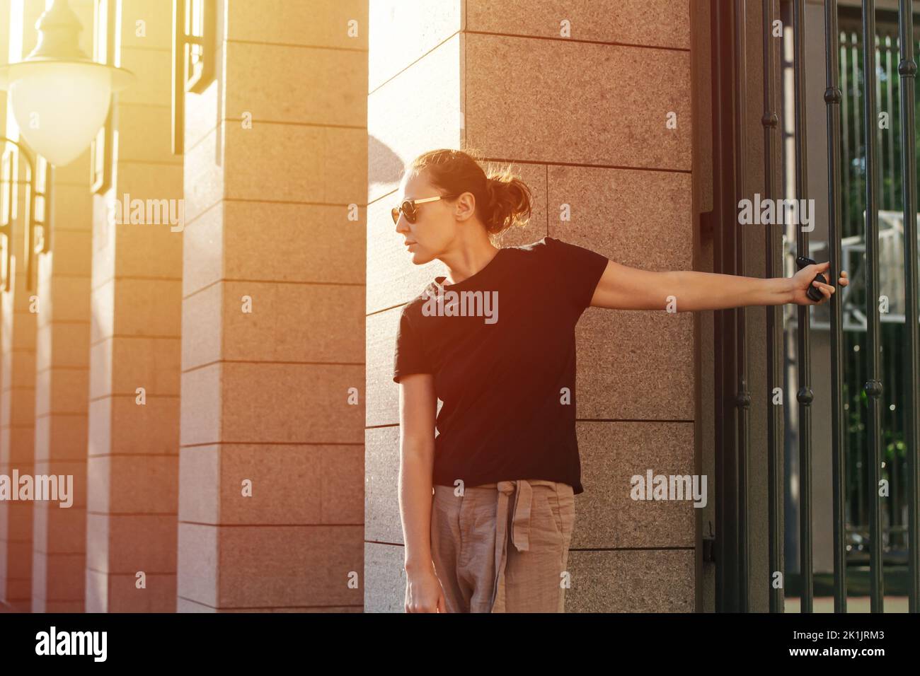 Profile image of a young woman in sunglasses holding on a fence rail behind her back on a sunlit street. Stock Photo