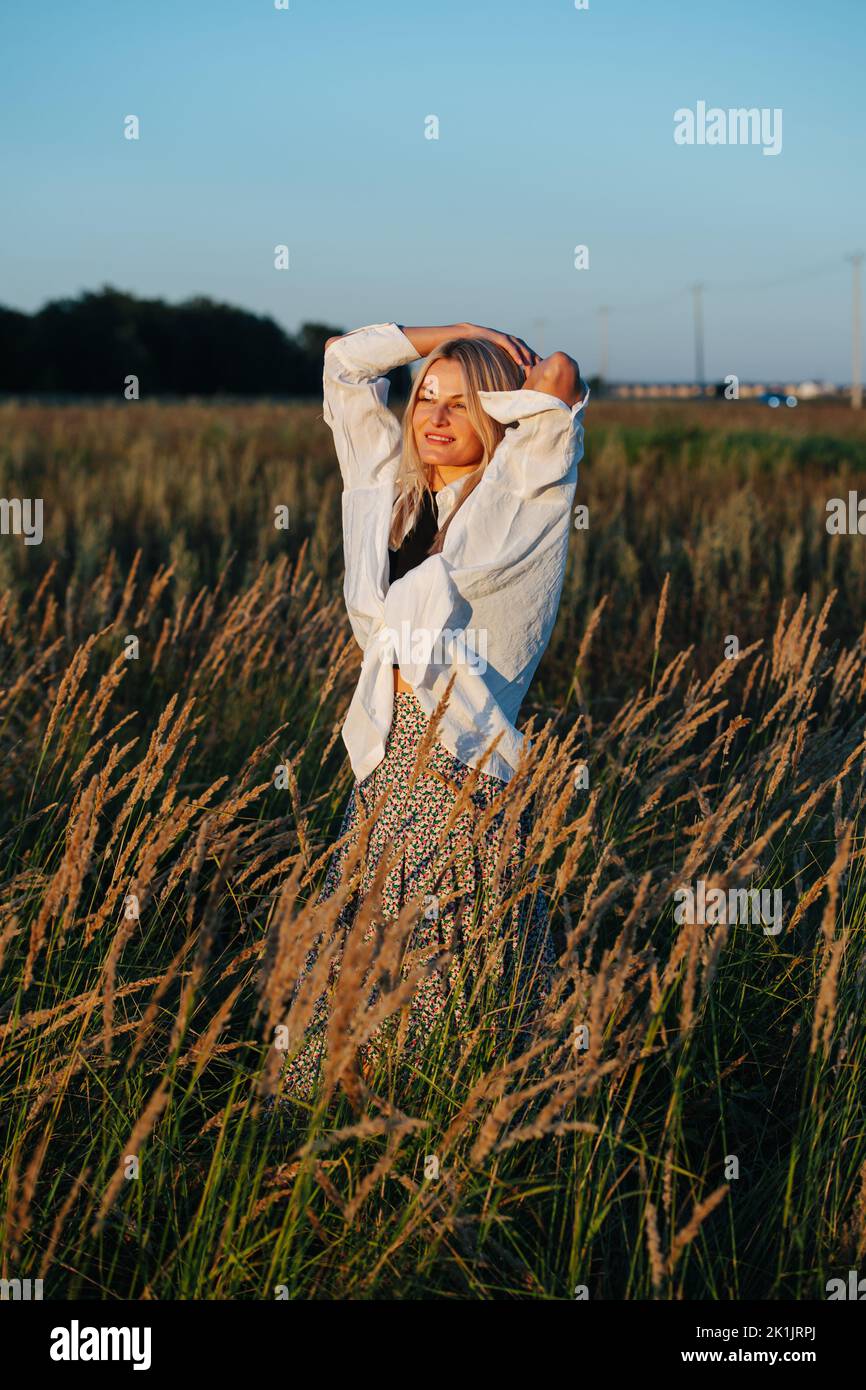 Peaceful young blond woman looking afar, standing amidst wheat field. Her hands on a back of her head. Blurred background, sharp foreground. Stock Photo