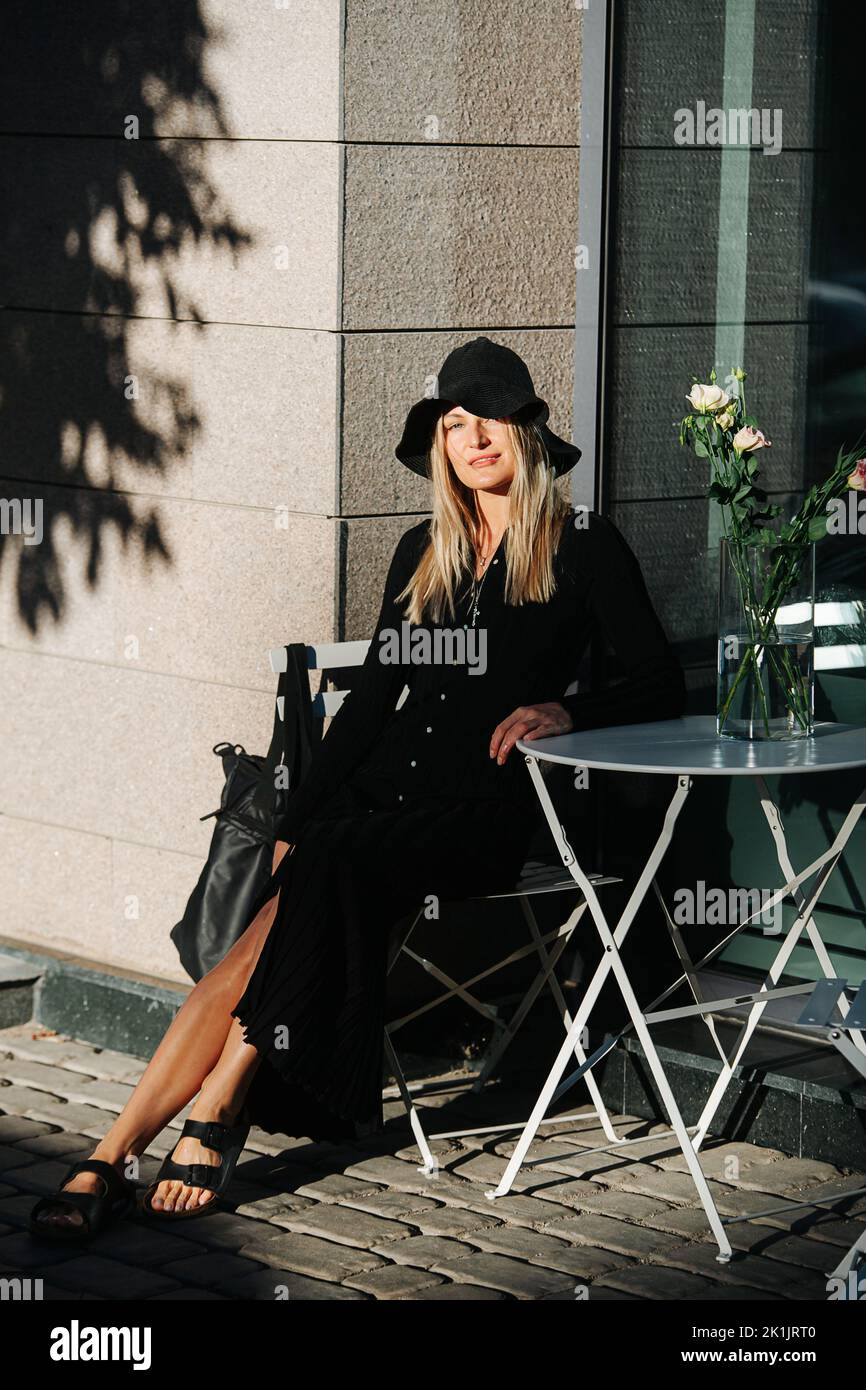 Covered young blond woman in floppy hat and long sleeved balck dress, sitting behind cafe table outside, looking at the camera. Stock Photo