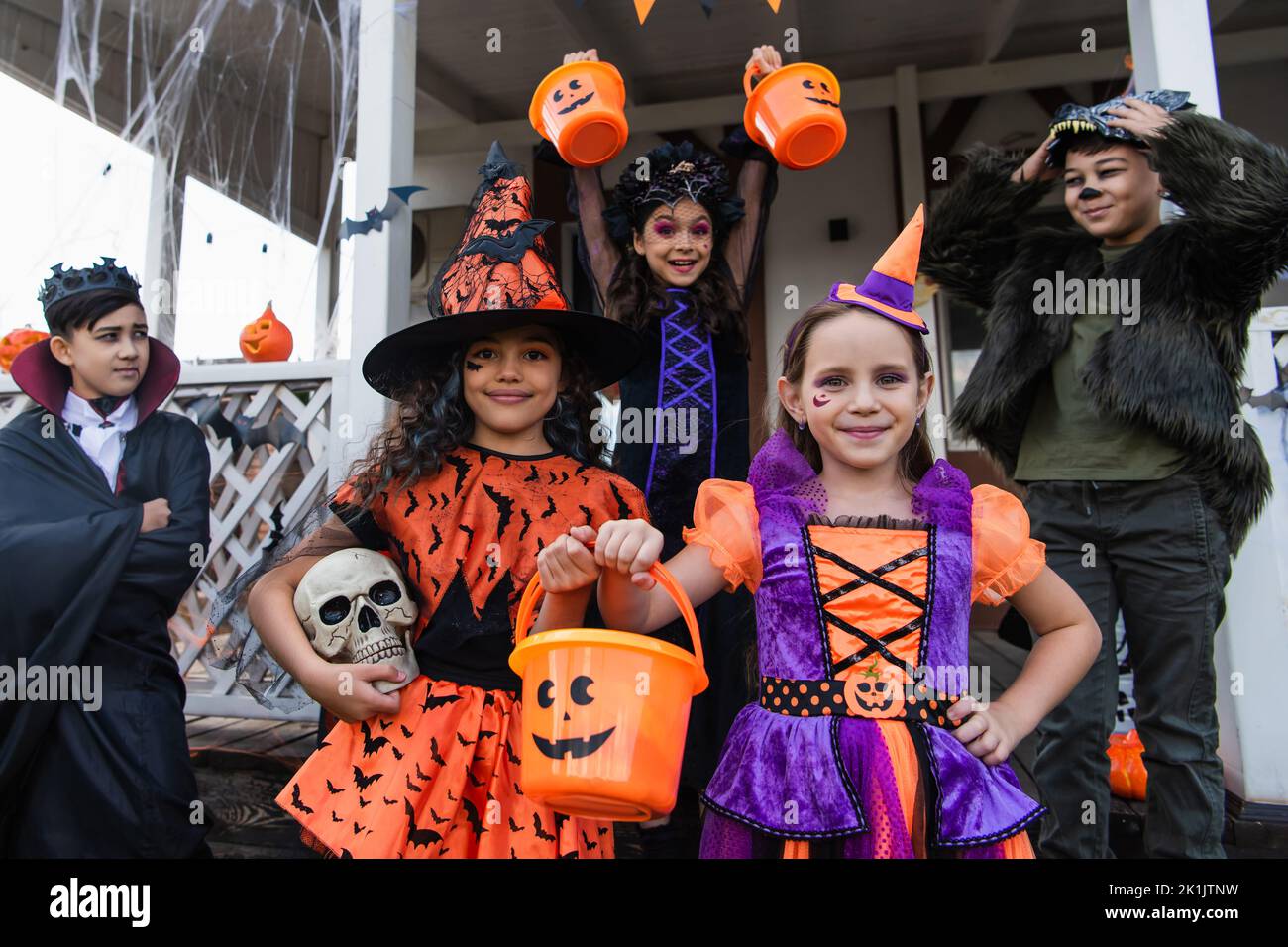 cheerful interracial kids in halloween costumes standing with trick or ...