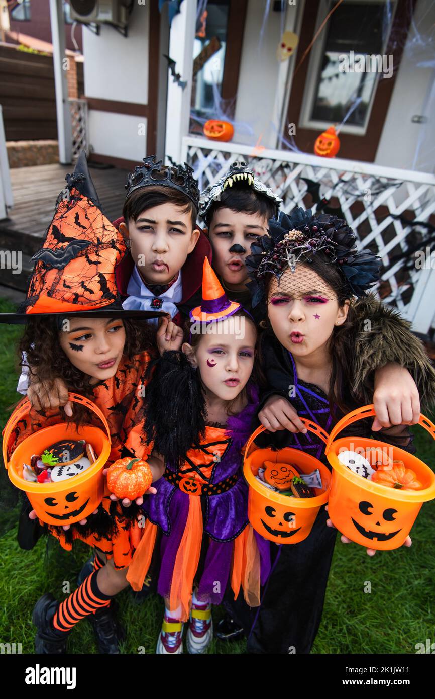 Multiethnic kids in costumes holding buckets with candies and grimacing ...