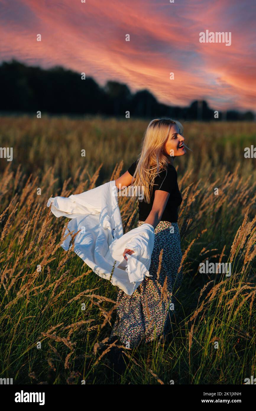 Carefree young blond woman spinning in a wheat field. Her shirt is falling off of her arms. Blurred background. Beautiful evening sky. Stock Photo