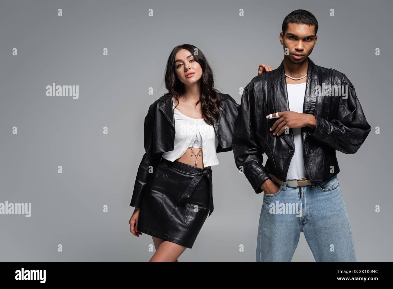 stylish african american man posing with hand in pocket near young woman in leather outfit isolated on grey,stock image Stock Photo