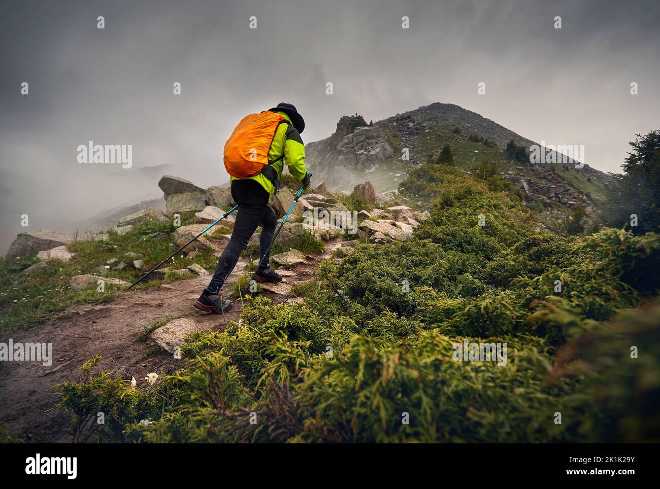Man Hiker tourist with orange backpack walk on the rock hill against fog and dark overcast sky in the mountain valley. Outdoor and trekking concept. Stock Photo