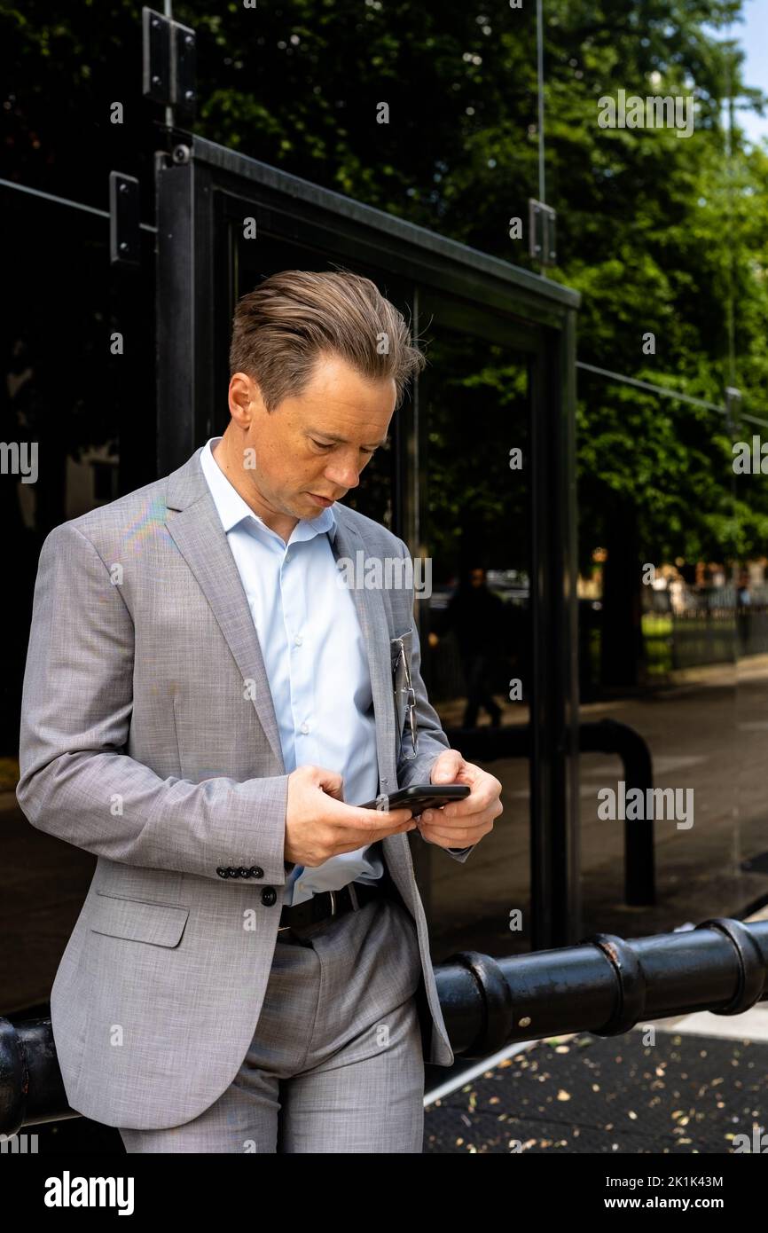 A handsome businessman in a grey suit standing outside his office while using his mobile phone. Working lunch concept Stock Photo