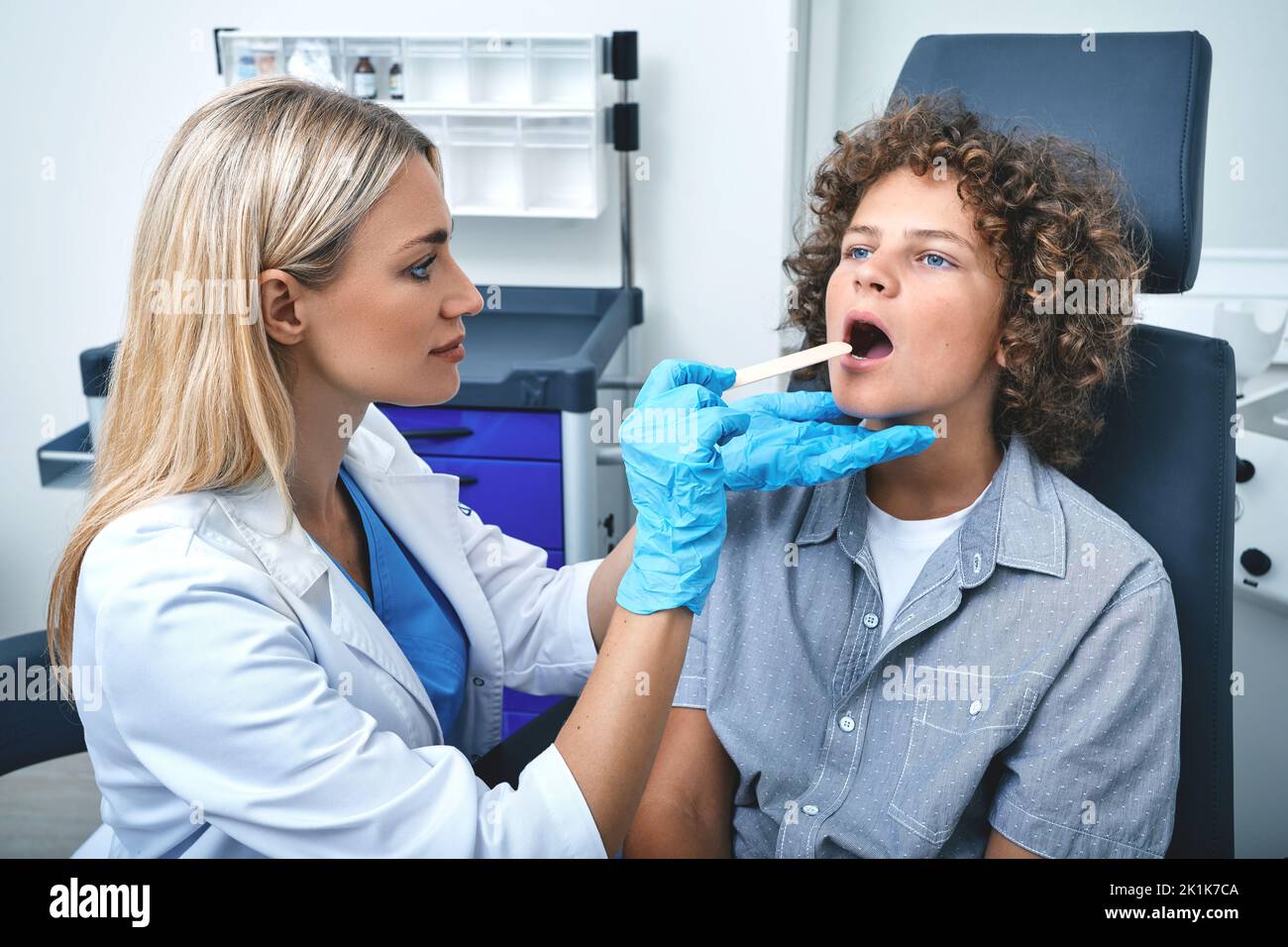 Otolaryngologist using inspection spatula to examines throat of male child. Curly boy patient opens his mouth during throat check-up Stock Photo