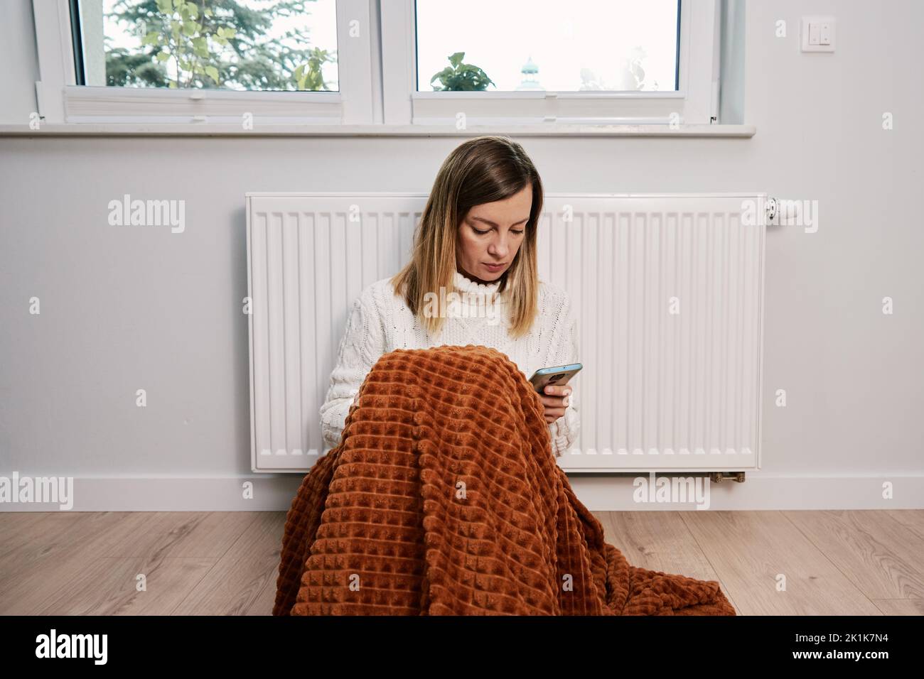 Worried sad woman sits under blanket near heating radiator and use smartphone, Rising costs in private households for gas bill due to inflation and war, Energy crisis Stock Photo