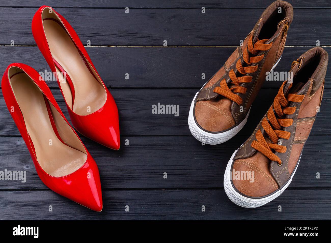 Red female high heel shoes and brown male sneakers. Top view mens and womens shoes on dark wooden background. Stock Photo