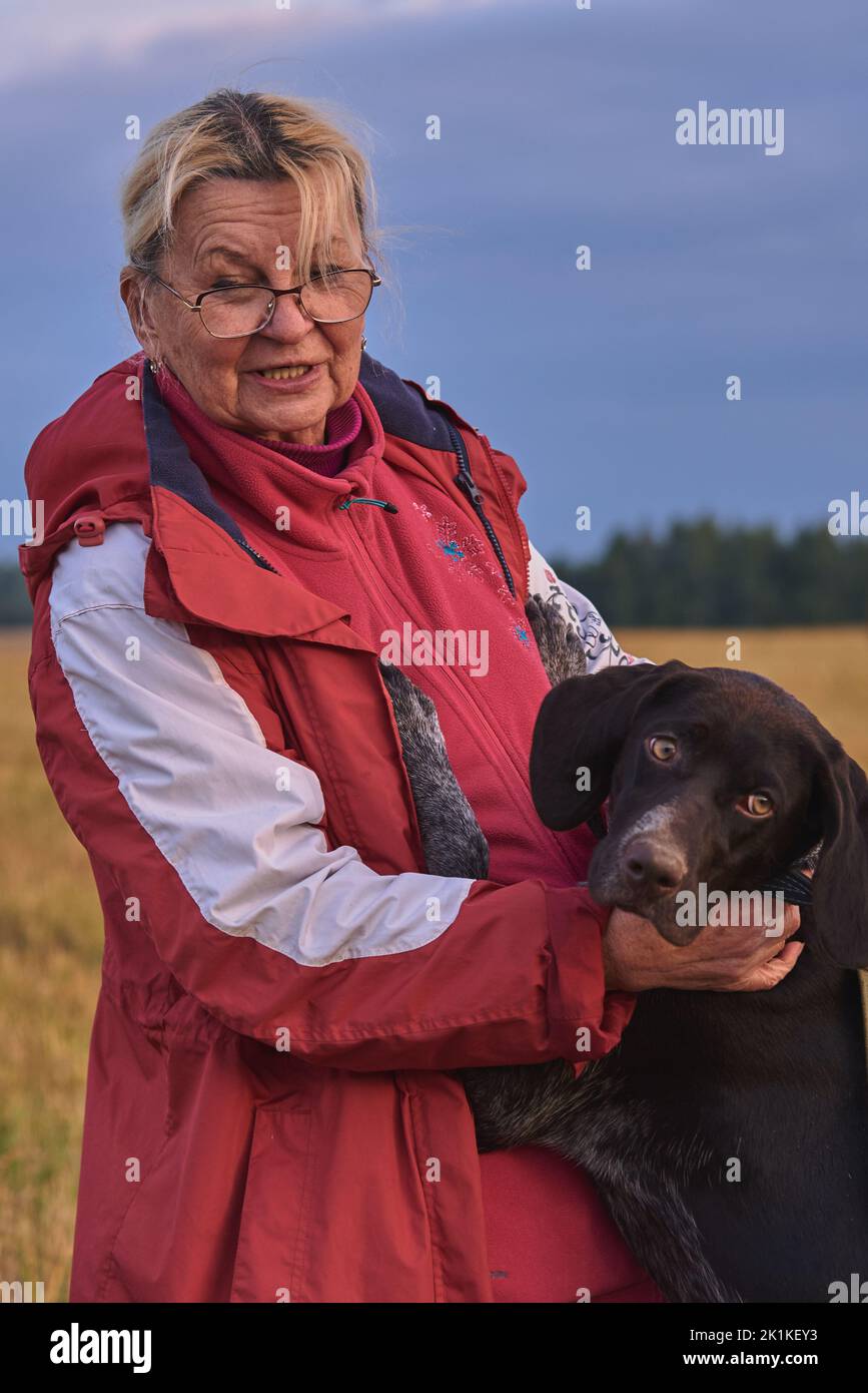 An elderly woman hugs her dog , German shorthaired pointer , in a mown field at sunset. Active longevity, a symbol of friendship and loyalty. Stock Photo