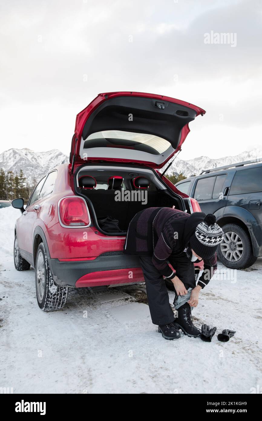 Woman putting on ski boots at back of car in snowy parking lot Stock