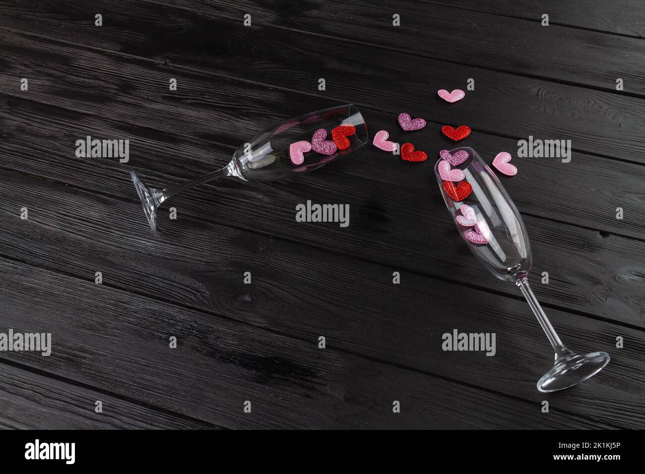 Two champagne glasses with spilled sequin hearts. Black wooden desk. Happy valentines day flat lay. Stock Photo