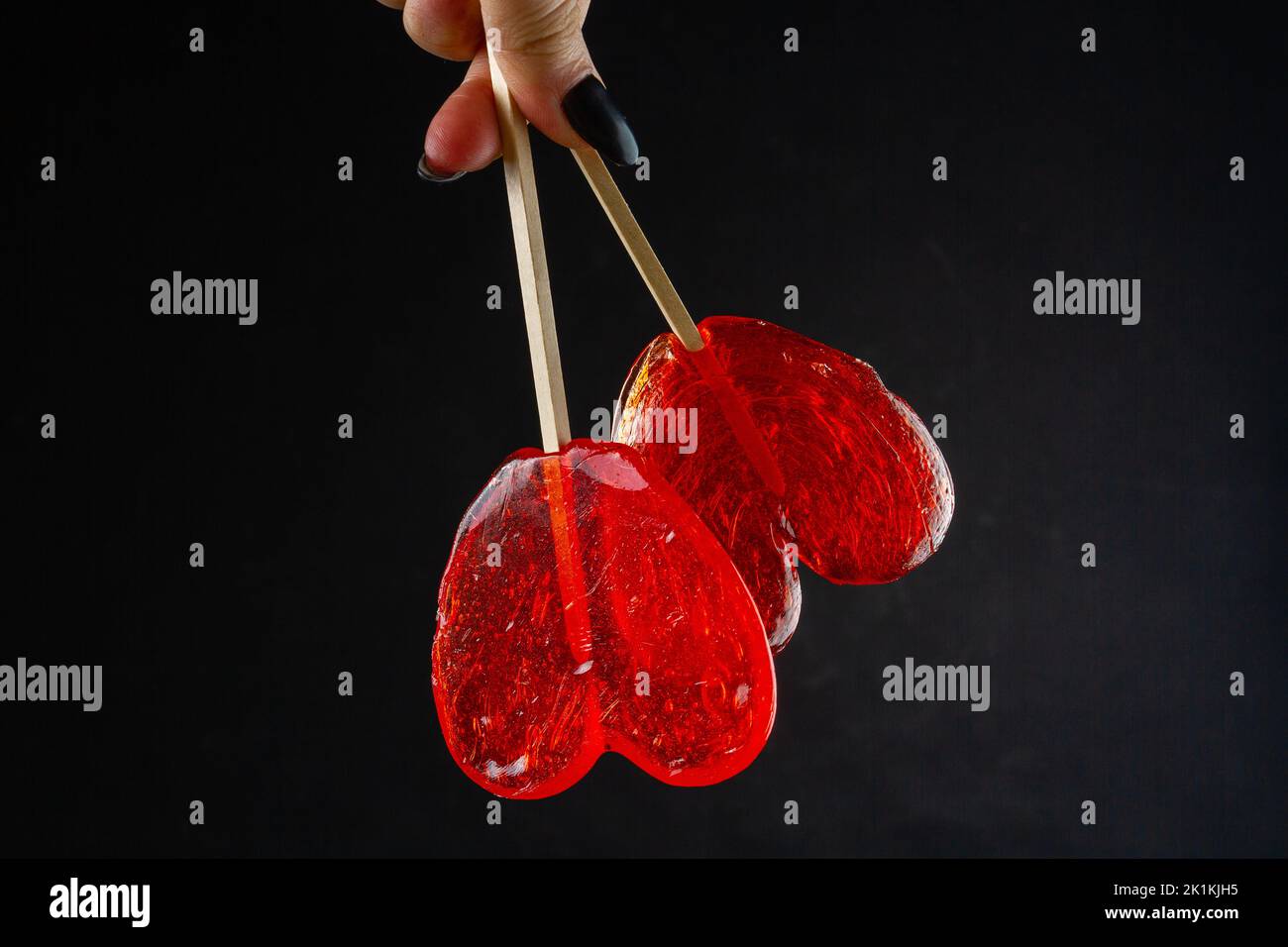 Hand holding red heart candies on black background. Pair of sweet lollipops. Stock Photo