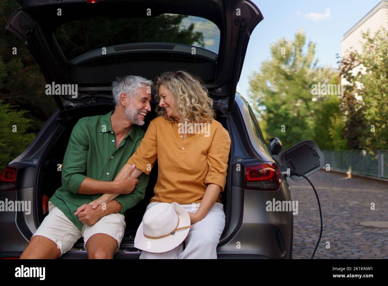 Middle-aged couple sitting in trunk while waiting for charging car before travelling on summer holiday. Stock Photo