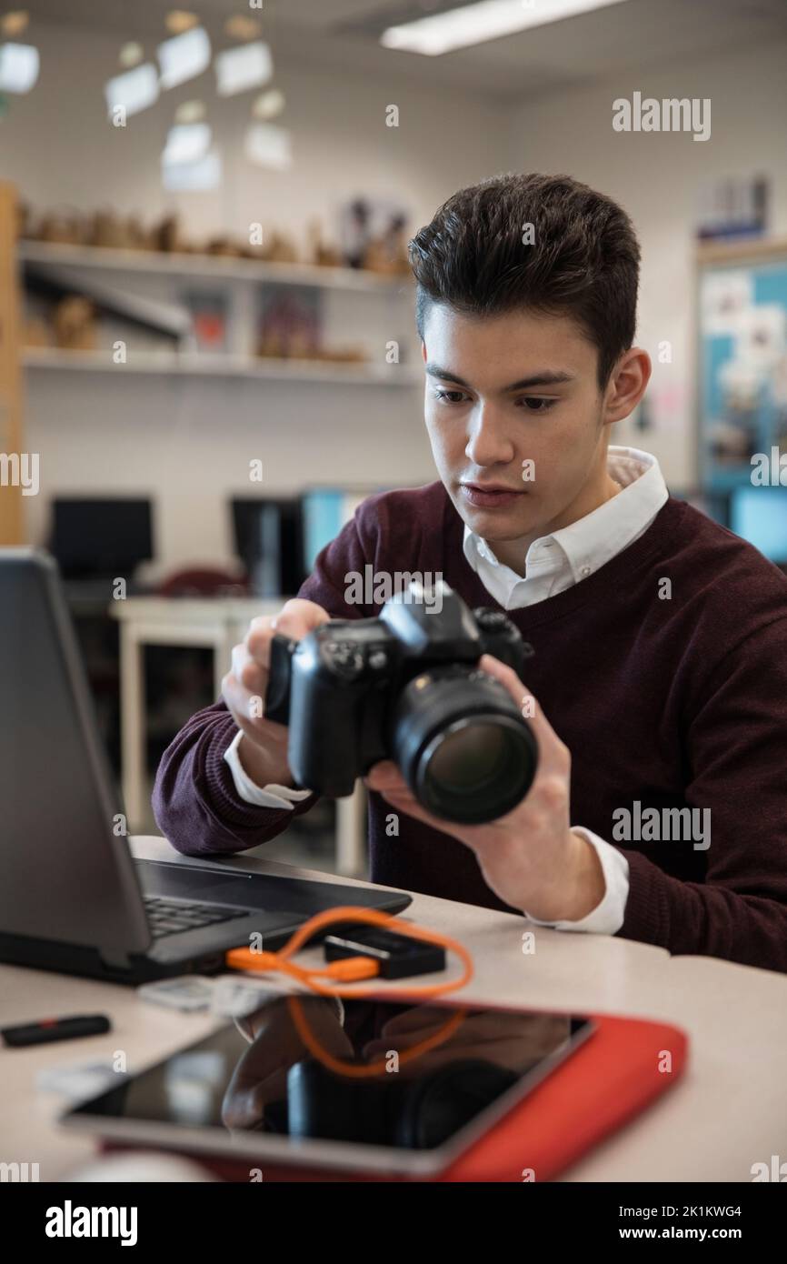 Focused high school boy using digital camera at laptop in classroom