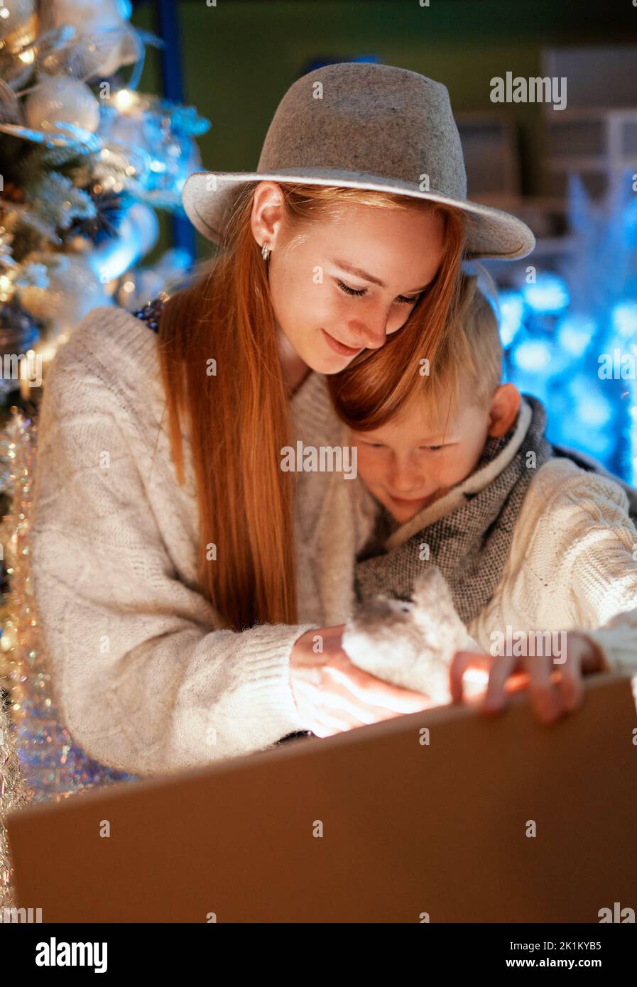 New Year 2023: Adorable brother and sister opens Christmas gift at home with xmas tree on background. Happy emotions, laughing while opening a present with light inside. High quality vertical image Stock Photo
