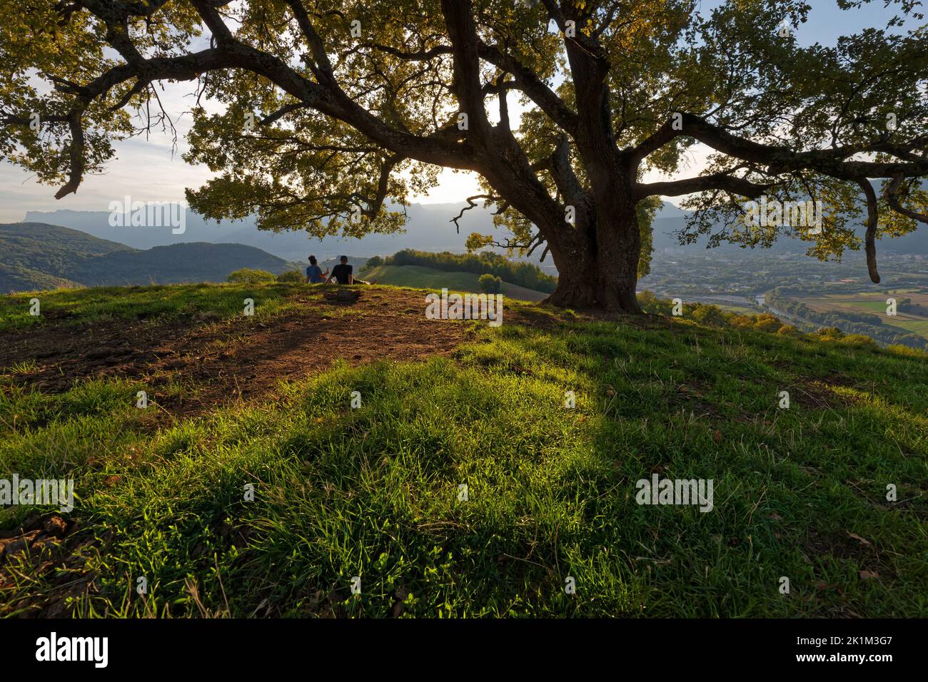 VENON, FRANCE, September 1, 2022 : Young couple rest at sunset under an old oak, called 'Le Chene de Venon', one of famous tree in french alps. Stock Photo