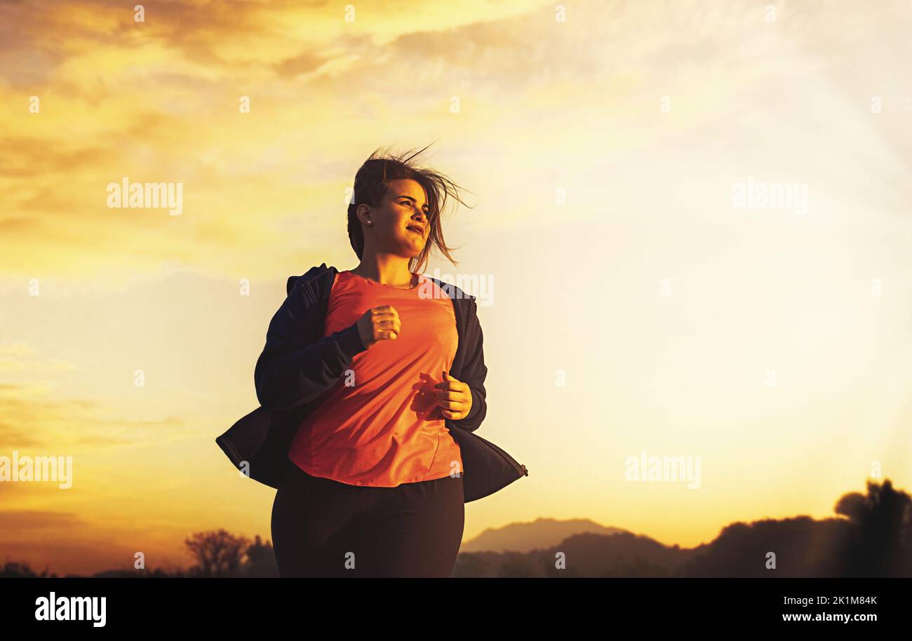 A beautiful plus size young woman jogging in the nature at sunset or sunrise determinate loose weight - people, sport, body positive and weight loss l Stock Photo