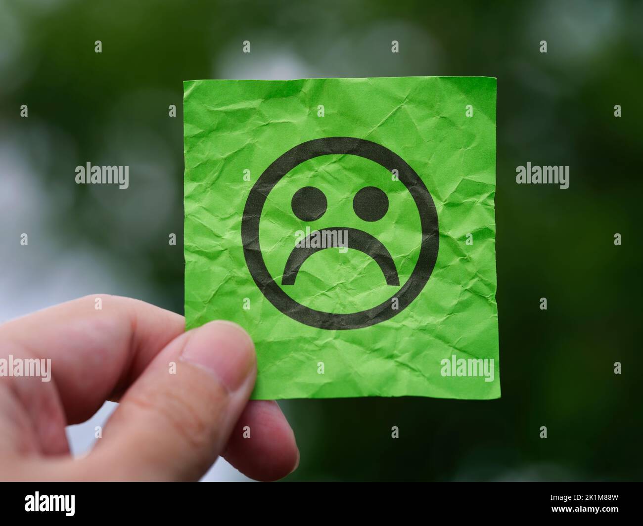 A man holding a green note paper with a sad face on it in his hand against a background of foliage. Close up. Stock Photo