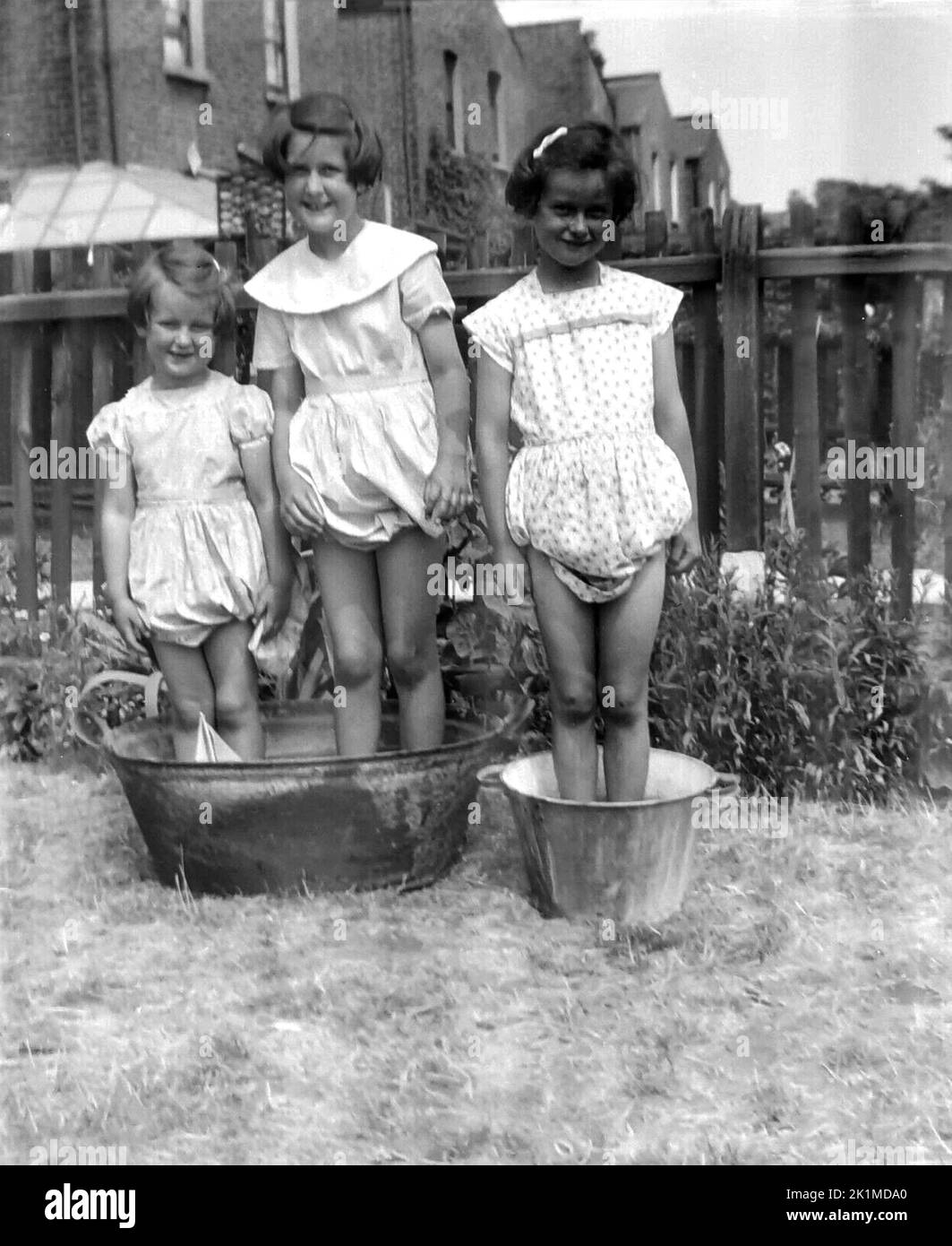 1940s, historical, three young girls playing outside, standing in two metal buckets in a back garden of a terraced house, England, UK. One of the buckets is a traditonal galvanised tin wash tub common for laundry and washing clothes in this era. Stock Photo