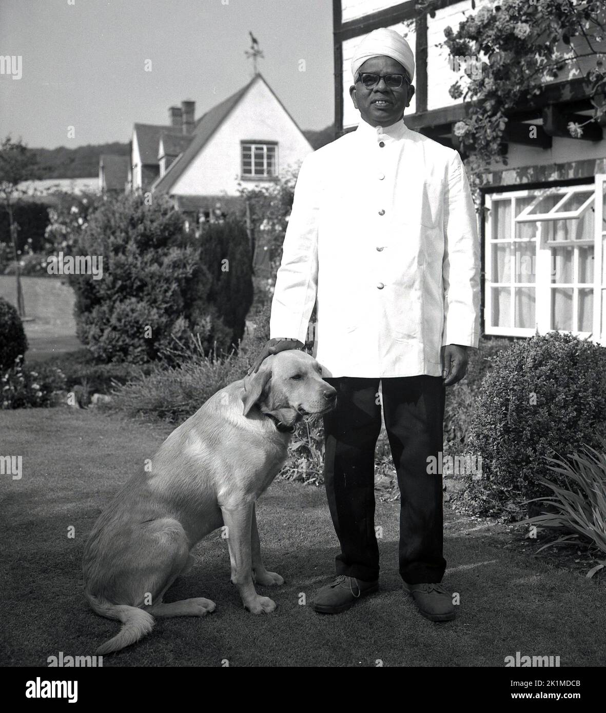 1950s, historical, a household servant, possibly a man friday or butler, an indian man in a uniform, a white tunic and wearing turban standing for a photo with the family pet dog, England, UK. Olinks??? Stock Photo