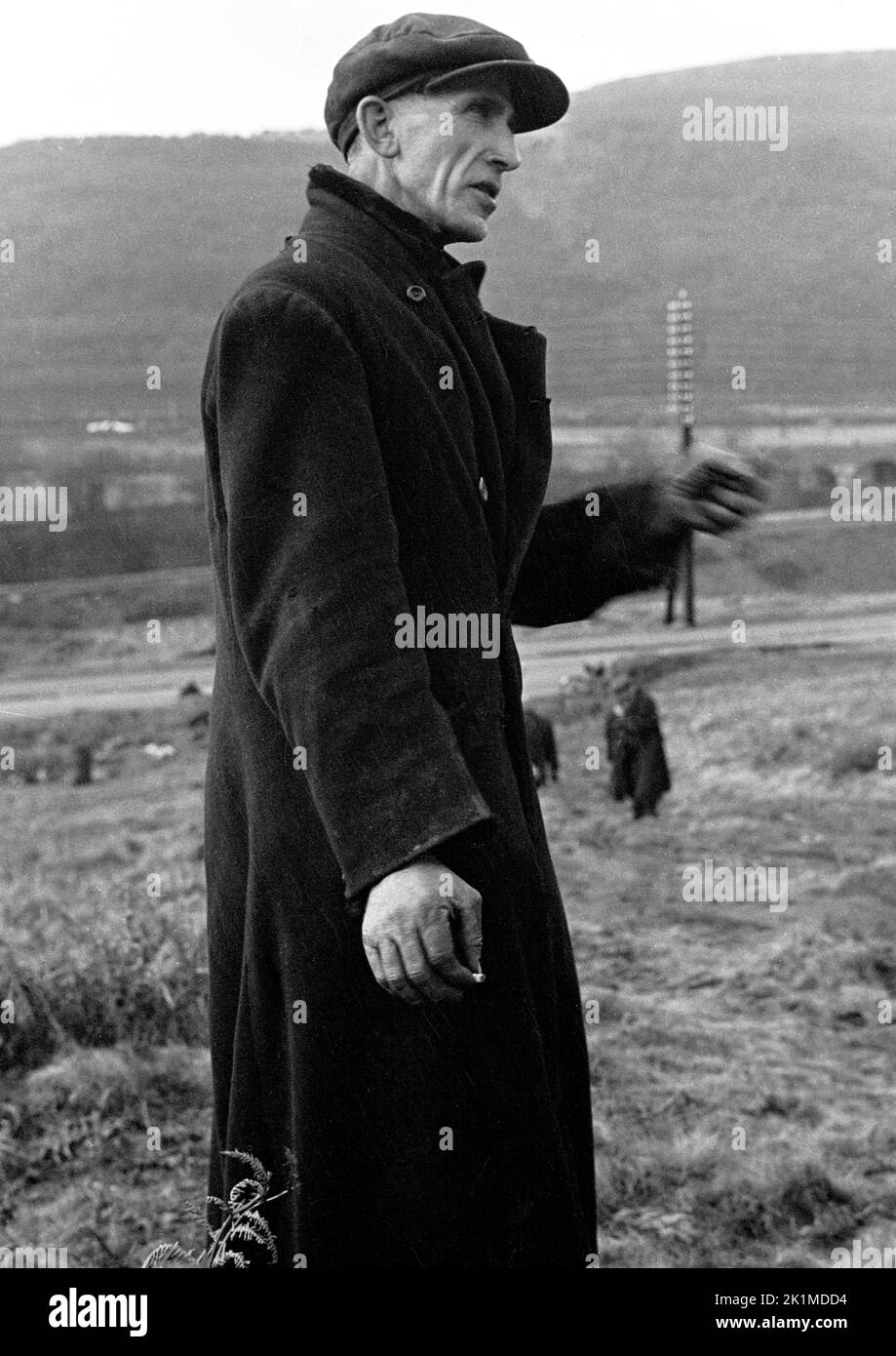 1930s, historical, a former Welsh mine worker in a flat cap and wearing a ragged patched coat standing on rough ground at a slag heap, Merthyr, South Wales, UK. Stock Photo