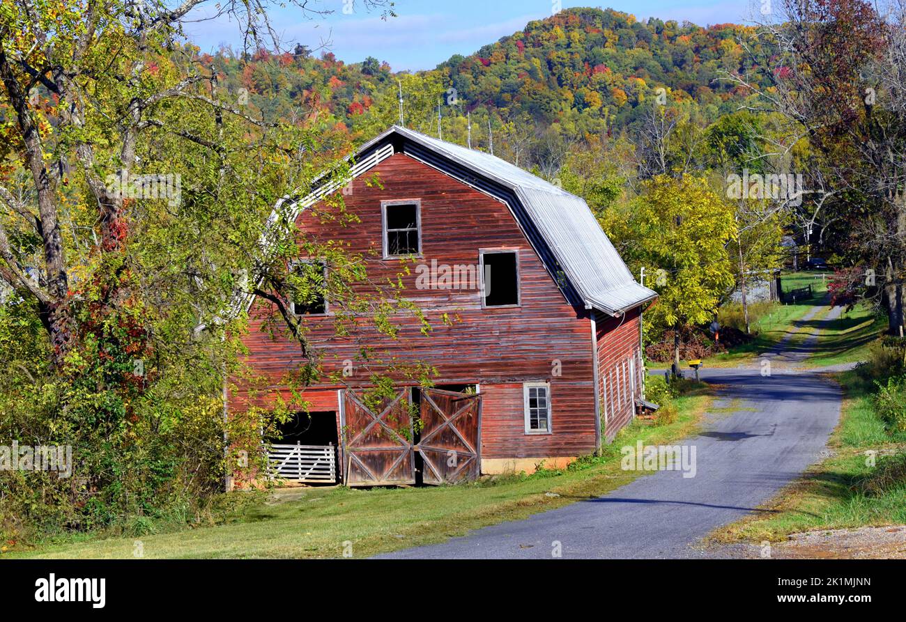 Rustic, red, wooden barn sits besides a narrow country lane.  Barn has tin roof and a hanging door. Stock Photo