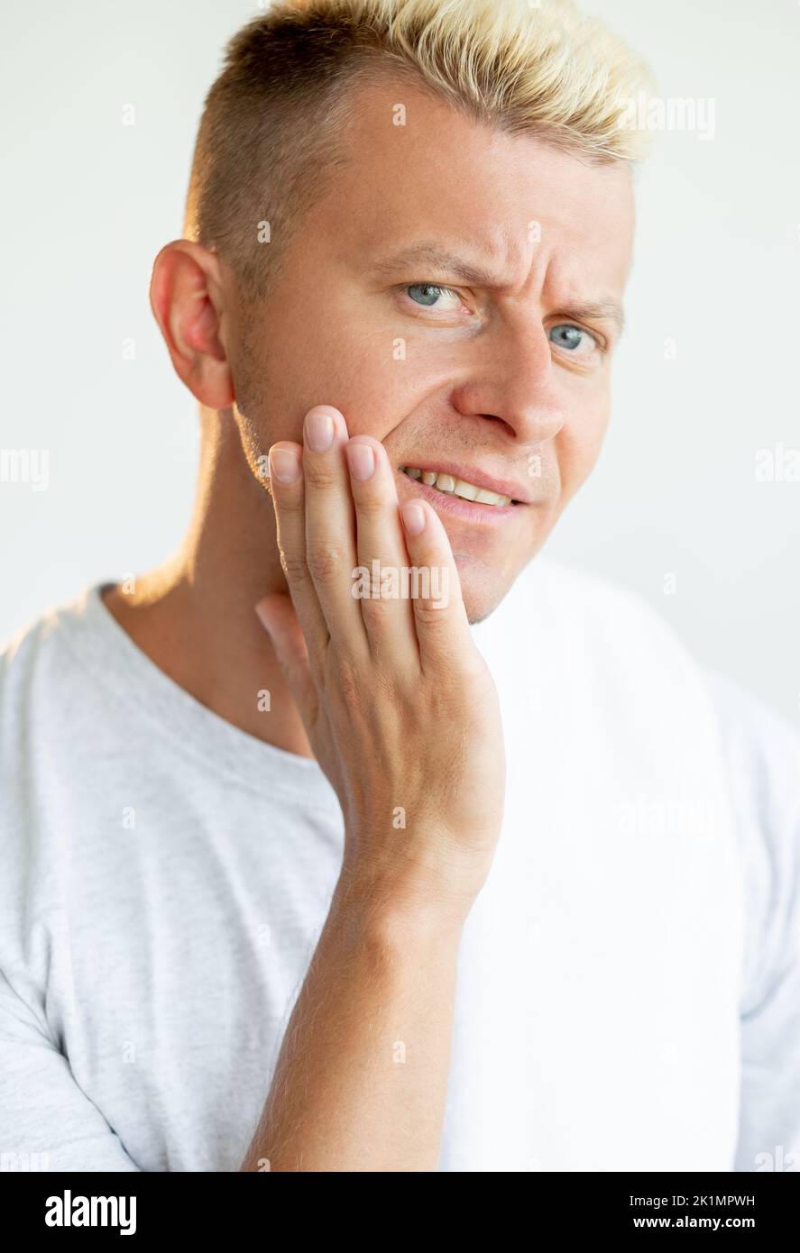 Male facial care. After shave skin moisturizing. Morning routine. Portrait of disturbed blond man in white t-shirt touching dry cheek isolated on ligh Stock Photo