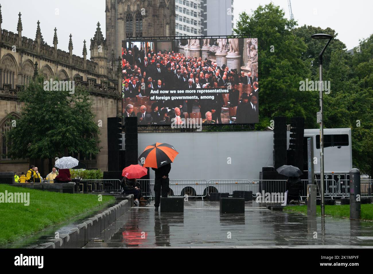 Electronic display screen in Cathedral Gardens showing funeral of monarch Queen Elizabeth II. People watching in rain with umbrellas. Stock Photo