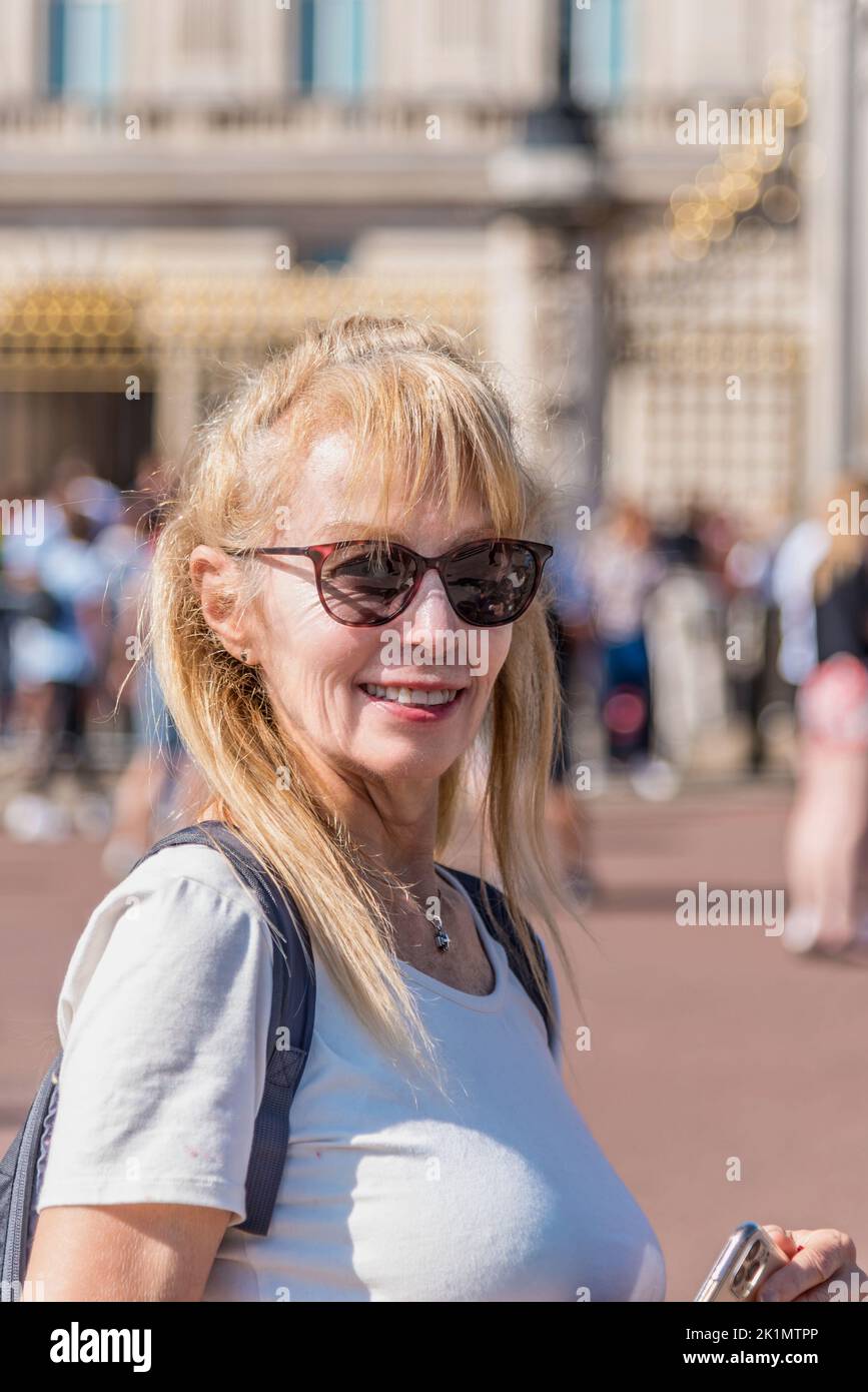 Portrait of a mature blonde woman with sunglasses looking at the camera. Unfocused Buckingham Palace and people behind. Stock Photo
