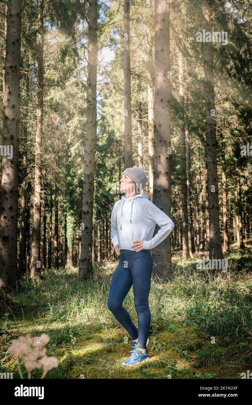 Sporty woman standing with her hands on her hips, resting after running in woodland. Stock Photo