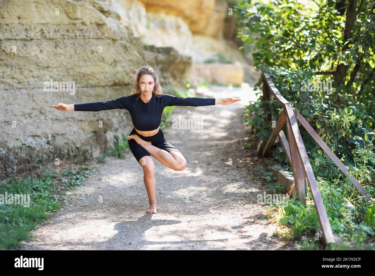 A woman in black sportswear practising yoga performs a variation of a vrikshasana exercise, a tree pose, trains in the park on a warm summer morning, Stock Photo