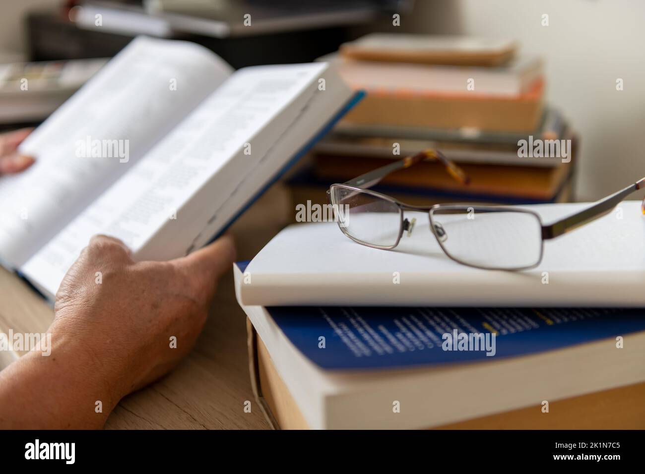 A pair of glasses on a desk with a person reading a book. Stock Photo