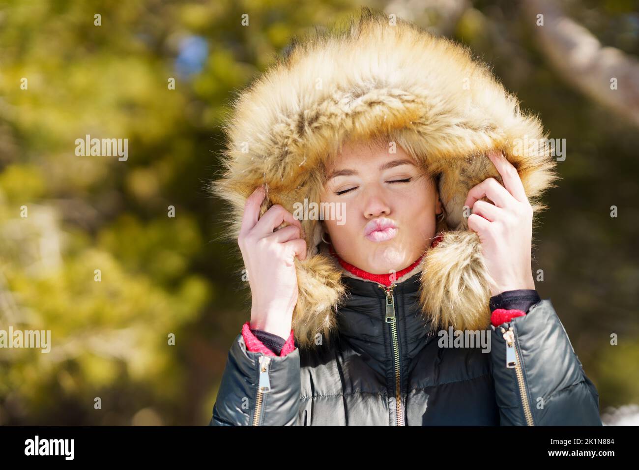 Young woman wearing a furry hooded jacket blowing a kiss in a forest. Stock Photo