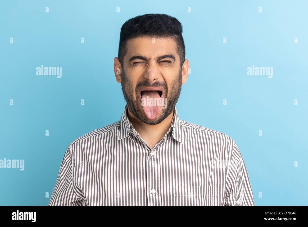 Photo of young handsome foolish crazy businessman, keeping eyes closed and showing tongue out, expressing positive emotions, wearing striped shirt. Indoor studio shot isolated on blue background. Stock Photo