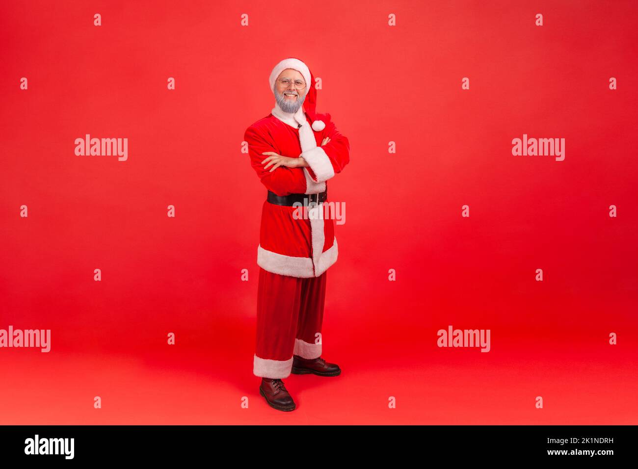 Full length photo of elderly man with gray beard wearing santa claus costume with confident look, standing with folded arms and looking at camera. Indoor studio shot isolated on red background. Stock Photo