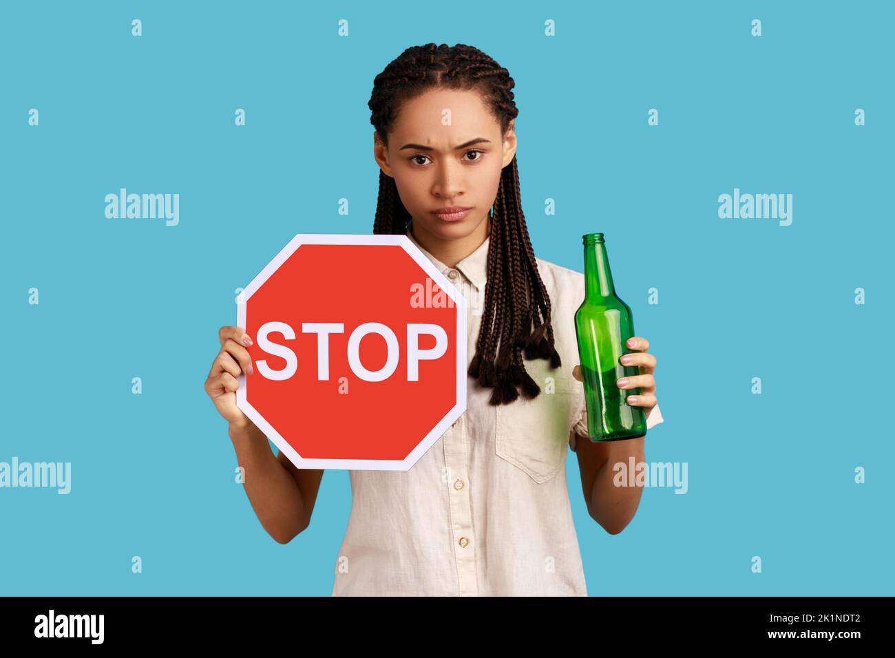 Portrait of serious woman with dreadlocks holding red stop sign and bottle with alcoholic beverage, calls on dont drink alcohol, wearing white shirt. Indoor studio shot isolated on blue background. Stock Photo