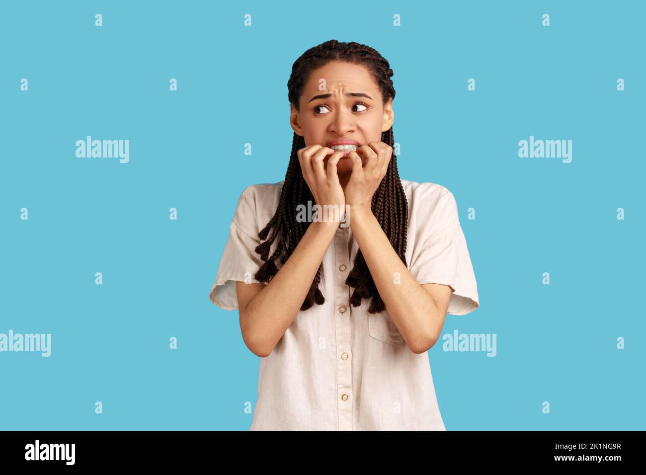 Portrait of puzzled woman with black dreadlocks has worried nervous expression, keeps hand on mouth, hears unexpected bad news, wearing white shirt. Indoor studio shot isolated on blue background. Stock Photo